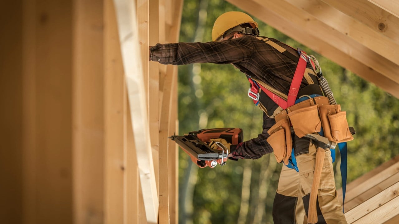Travailleur en construction portant un casque et un harnais de sécurité, utilisant une cloueuse pneumatique sur une charpente en bois.
