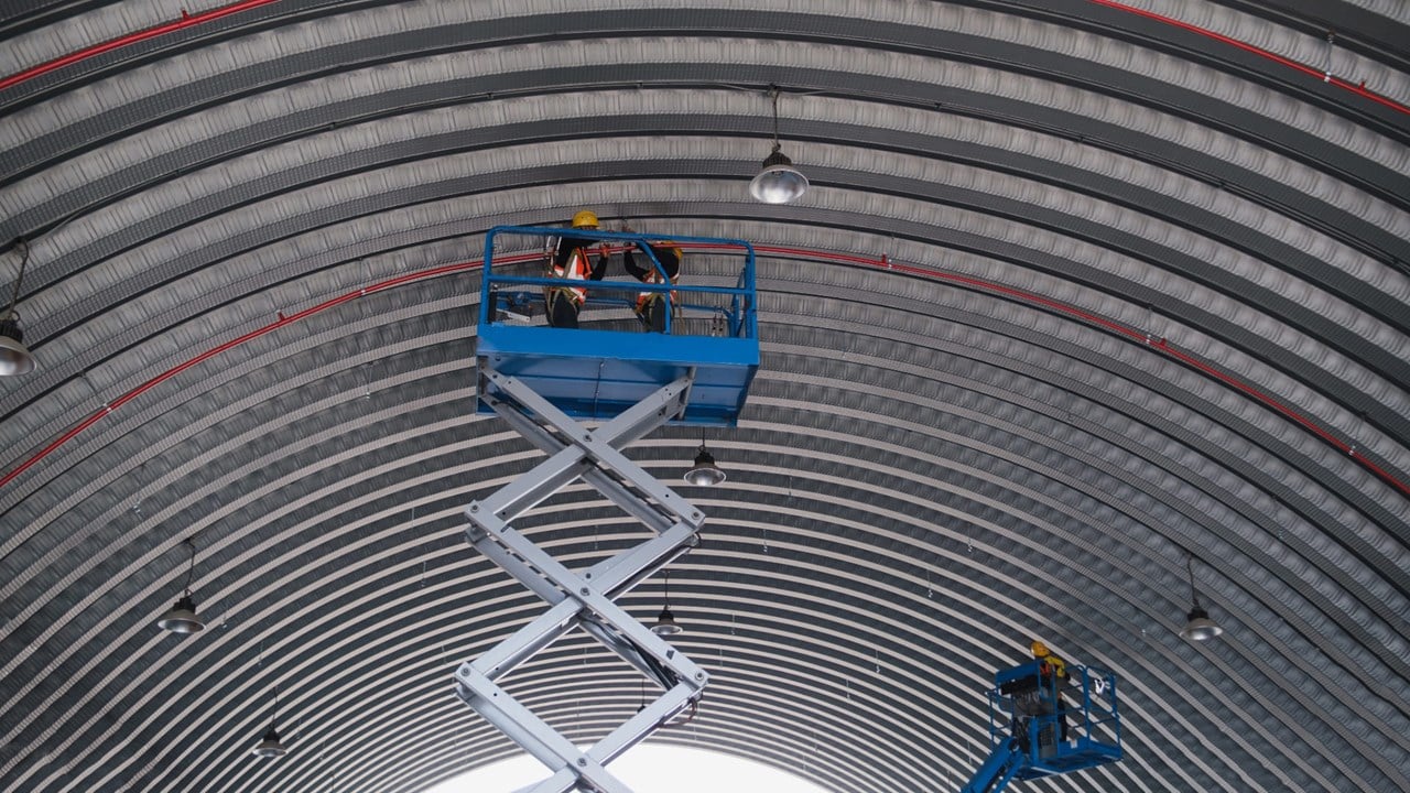 Travailleurs portant un casque et un harnais de sécurité sur une plateforme élévatrice à ciseaux et une nacelle, installant des structures en hauteur.