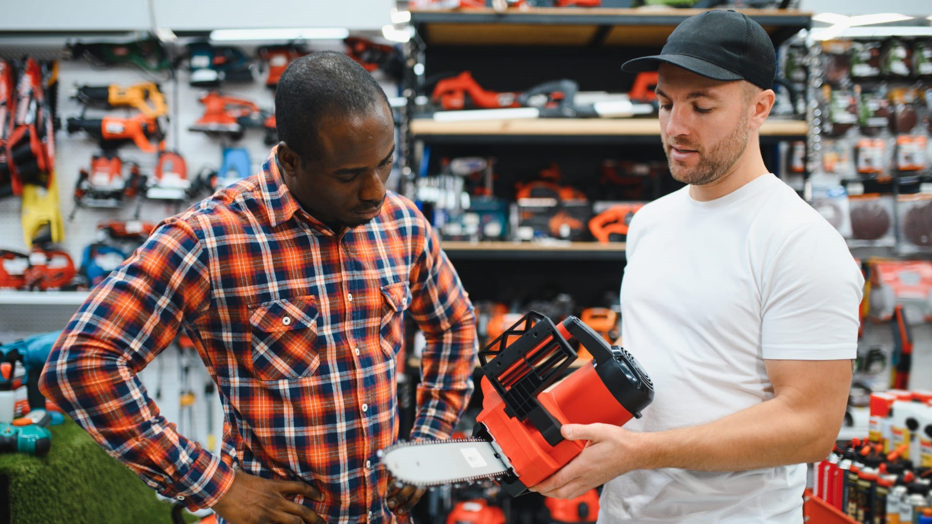 Un employé Simplex en t-shirt montre une tronçonneuse à un client en chemise à carreaux, devant un mur de perceuses et d’outils en magasin.