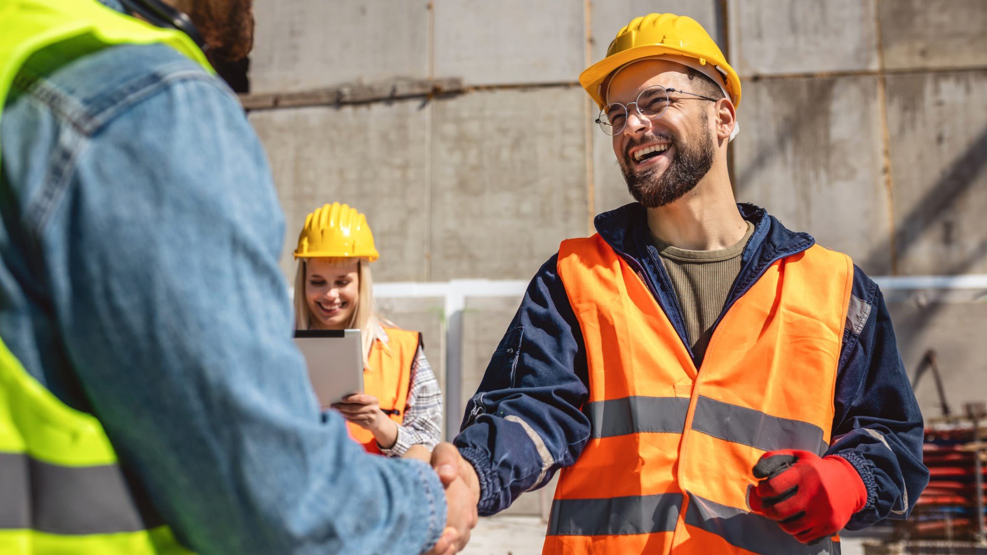 Un ouvrier avec un casque de chantier souriant, serre la main à une autre personne de dos. Une femme avec un casque de chantier est en arrière plan.