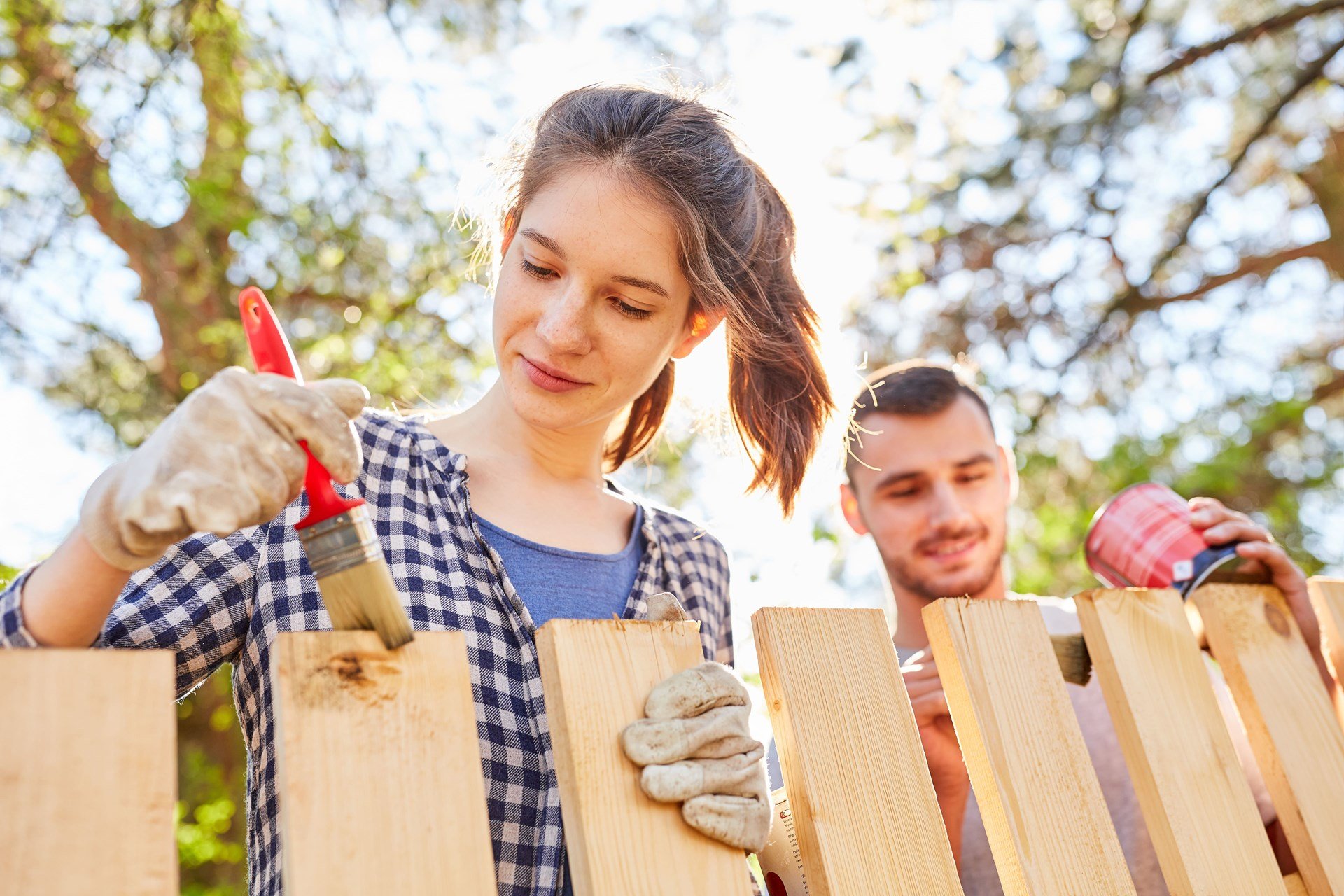 Femme peignant une clôture en bois avec un pinceau rouge pendant qu’un homme prépare la teinture, dans un jardin en été